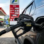 Foto close-up dramatis dari nosel pengisian BBM jenis Dexlite atau Pertamina Dex yang sedang mengalirkan bahan bakar ke tangki mobil SUV modern. Latar belakang memperlihatkan papan digital harga SPBU Pertamina yang menyala terang, diambil dengan sudut estetik (low-angle) saat senja untuk memicu perhatian pengguna HP.