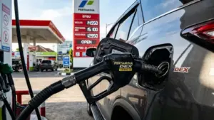 Foto close-up dramatis dari nosel pengisian BBM jenis Dexlite atau Pertamina Dex yang sedang mengalirkan bahan bakar ke tangki mobil SUV modern. Latar belakang memperlihatkan papan digital harga SPBU Pertamina yang menyala terang, diambil dengan sudut estetik (low-angle) saat senja untuk memicu perhatian pengguna HP.